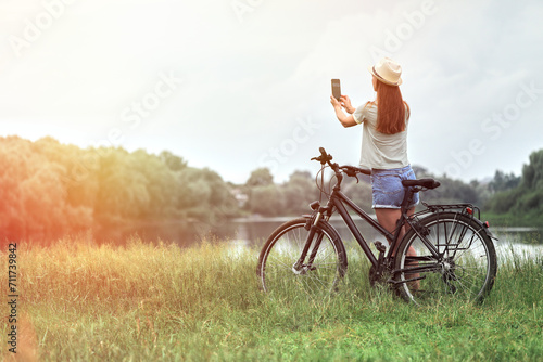 Woman walking with bicycle using mobile phone in park or field. ECO friendly transport concept. Cyclist stopped by the road to look at her cell phone.
