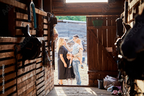 A happy family spends their leisure time at a farm with horses and other animals. Parents with children in the background of a stable or barn.