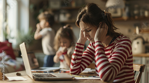 Stressed woman sitting at a table in front of a laptop, holding her head in her hands, with children in the background, reflecting the challenges of balancing work and childcare at home. photo