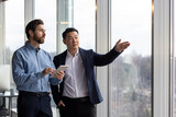 Two professional businessmen engaged in a discussion while one holds a tablet and the other points out of a window in a corporate office setting.