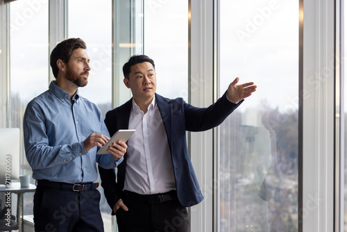 Two professional businessmen engaged in a discussion while one holds a tablet and the other points out of a window in a corporate office setting.