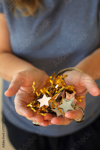 Middle-aged woman holding gold and silver star shapped birthday confetti in hands while standing against beige wall