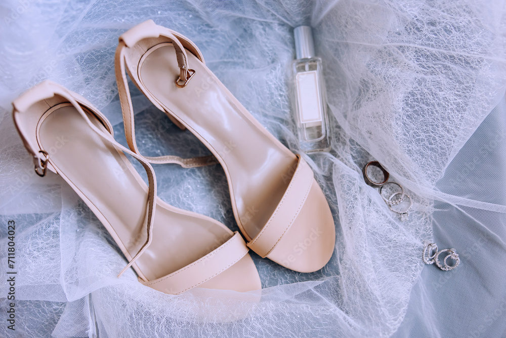 Wedding shoes for the bride. Elegant sandals of beige color for the bride stand on a white tulle. Wedding rings, earrings and perfumes. Bride's accessories. Wedding day Wedding shoes for the bride. Elegant sandals of beige color for the bride stand on a white tulle. Wedding rings, earrings and perfumes. Bride's accessories. Wedding day