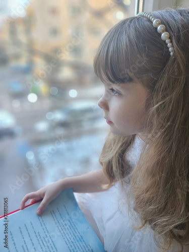 A cute child girl in white, holding a book, looking at the winter nature from the window, in her room at home