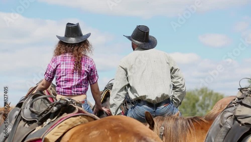 Man and woman sit on fence among horses looking at sky, rear view photo