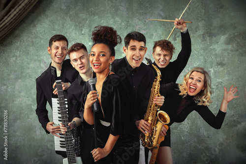 Cheerful international music group on a gray wall background, a group of musicians posing on camera in the hands of various instruments, guitars, saxophone. Copy space.
