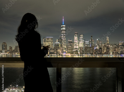 woman looking at downtown manhattan nyc skyline (after sunset, night time)  world trade center cityscape (high rise urban buildings) railing, hudson river, waterfront, bay, harbor new york city view