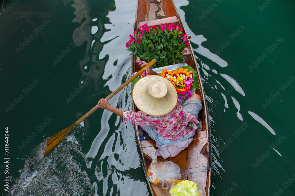 Fototapeta premium boater in Thailand shot from above, she is carrying flowers to the market and wearing a bamboo hat and holding an oar at the floating market Damnoen saduak near bangkok