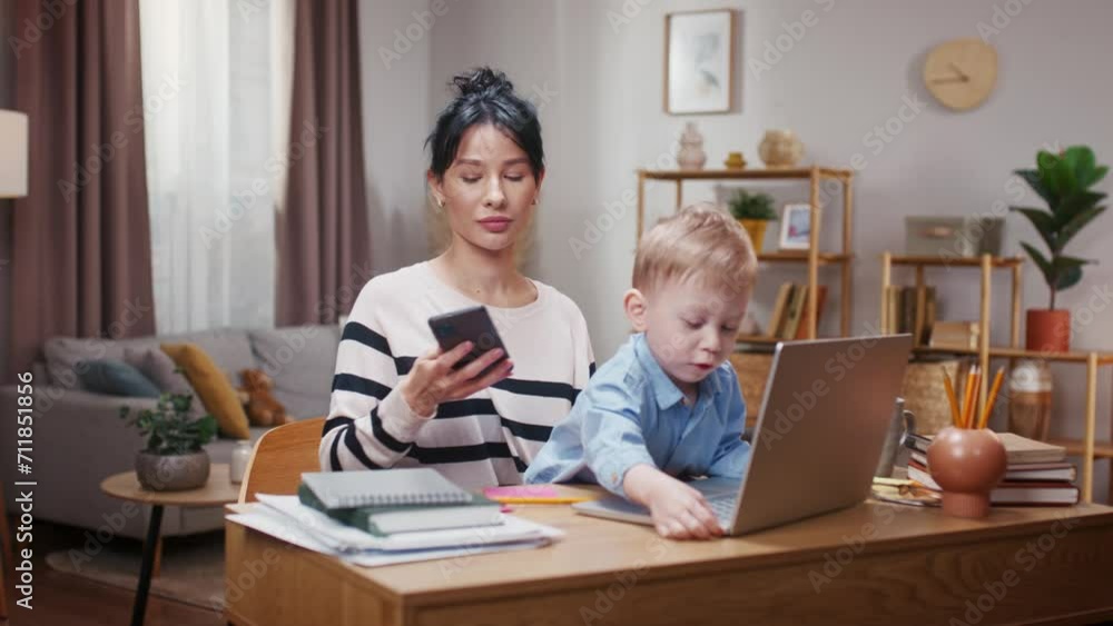 Woman working with smartphone and taking care of liitle son. Mother working distantly and babysitting with child. Boy clicking tablet keyboard and disturbing mother. Mom working online with kiddo. Woman working with smartphone and taking care of liitle son. Mother working distantly and babysitting with child. Boy clicking tablet keyboard and disturbing mother. Mom working online with kiddo.