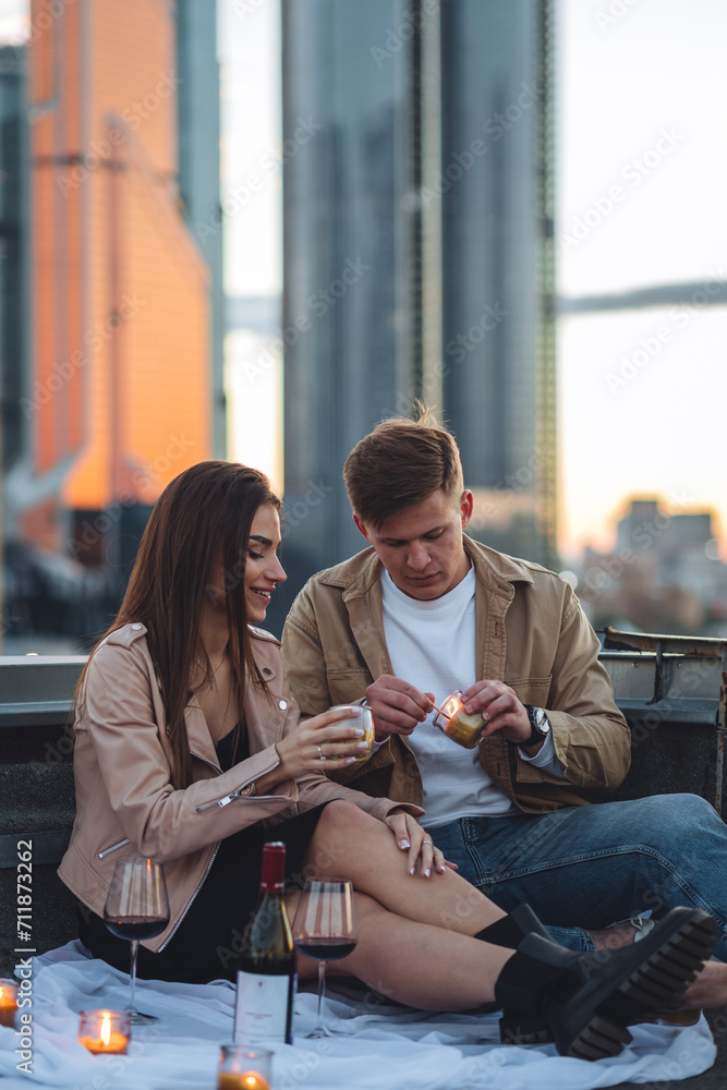 Fototapeta premium Romantic beautiful surprise on Saint Valentine's Day for a girlfriend. Loving young couple having dinner on a rooftop on sunset evening with candles, wine. Skyscrapers on background, urban cityscape
