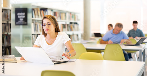 Wallpaper Mural Positive Latin young lady in casual clothes and glasses working remotely using a computer in a quiet library Torontodigital.ca