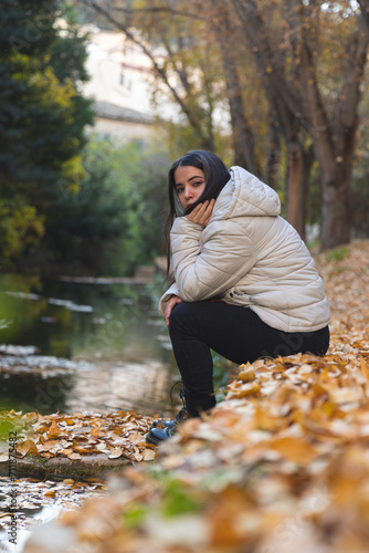 Mujer joven con chaqueta blanca expresa tristeza o relajación mientras se sienta junto a un lago rodeado de hojas de otoño