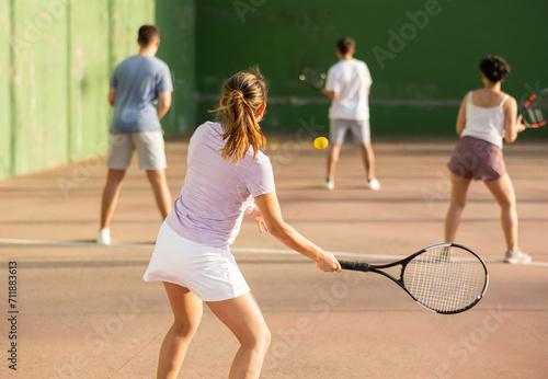 Rear view of young fit brown-haired woman playing frontenis team match on outdoor fronton court, swinging strung racket ready to hit small yellow rubber ball