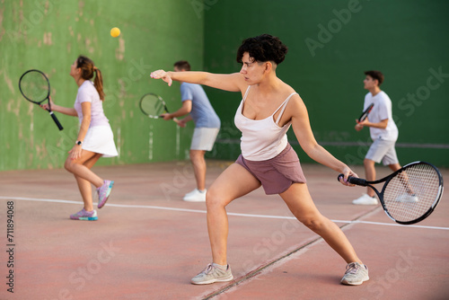 Concentrated young hispanic female frontenis player swinging string racquet to hit ball on outdoor three-walled court on sunny summer day. Sport and active lifestyle concept..