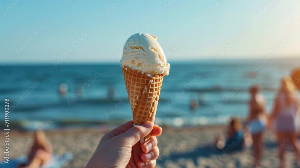 Fototapeta premium A person holds a vanilla gelato cone on a sunny beach with friends and family in the background, capturing a perfect summer day.