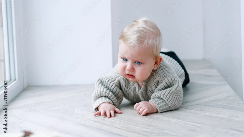 Adorable blond blue-eyed baby boy wearing grey sweater lies on his belly. Little kid near the window. Adorable blond blue-eyed baby boy wearing grey sweater lies on his belly. Little kid near the window.
