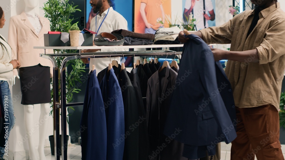 Man looking at elegant blazers in clothing store, trying to determine if materials are qualitative. Client analyzing garments on fashion boutique racks, deciding to buy after finding top quality Man looking at elegant blazers in clothing store, trying to determine if materials are qualitative. Client analyzing garments on fashion boutique racks, deciding to buy after finding top quality