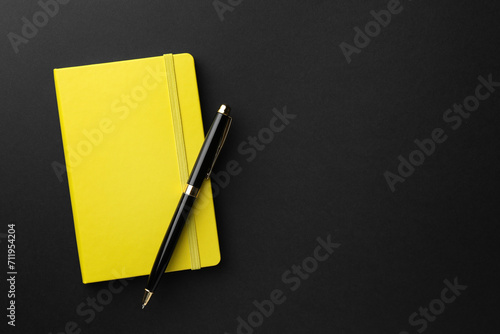 Closed yellow notebook and pen on black background, top view. Space for text