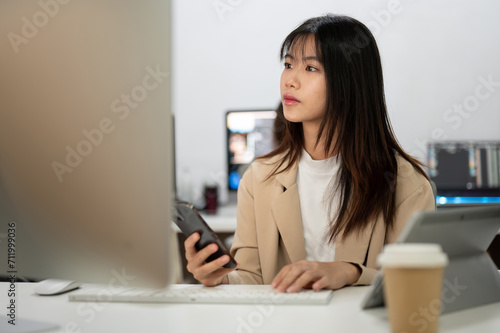 Young beautiful Asian businesswoman working in her agency office, using smartphone and computer. © bongkarn
