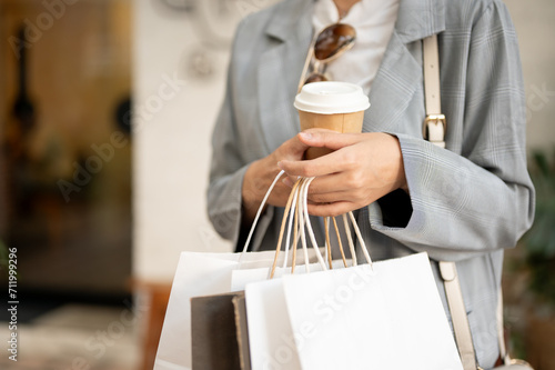Cropped image of a woman in trendy clothes carrying shopping bags and a coffee cup, walking outdoors