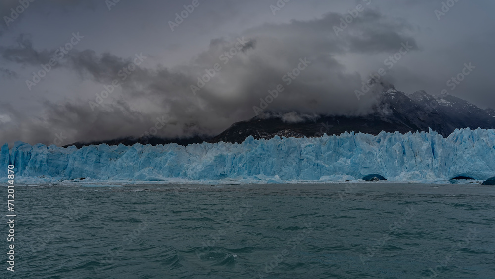 A wall of blue ice with cracks and sharp peaks stretches over a turquoise glacial lake. Melted ice floes float in the water. Mountains in clouds and fog. Perito Moreno glacier. El Calafate. 