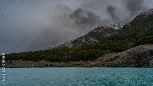 The beautiful turquoise glacial lake is surrounded by mountains. Coniferous forest grows on the rocky slopes. The layered structure of the cliffs is visible. Clouds, fog. Ripples on calm water.