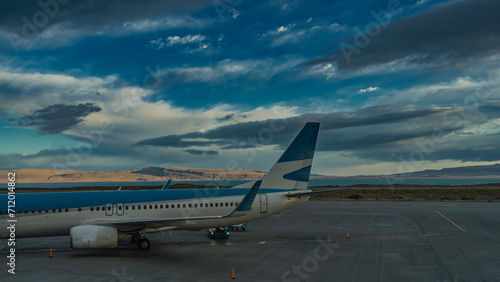 The plane is on the runway. There are marking strips and warning signs on the asphalt. In the distance there is a turquoise glacial lake, a mountain range against a blue sky and clouds. El Calafate.