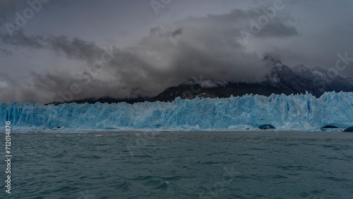 A wall of blue ice with cracks and sharp peaks stretches over a turquoise glacial lake. Melted ice floes float in the water. Mountains in clouds and fog. Perito Moreno glacier. El Calafate. 