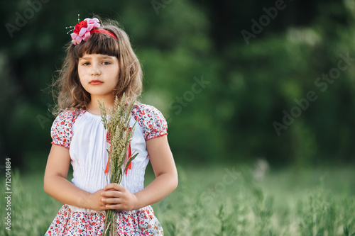 Little  beautiful and fragile Ukrainian girl on the background of nature with greens. Cute child portrait close-up with spikelet. Young child posing in colorful dress and bow.