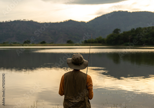 Enjoy moment of Handsome man fishing as a leisure activity during his vacation at the lake on sunset. Silhouette at sunset moment of man fishing rotation with reel. © Chanakon