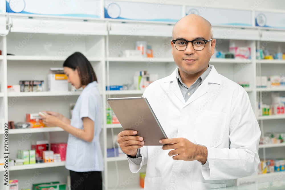Fototapeta premium Professional asian man pharmacist checks inventory arrangement of medicine in pharmacy drugstore. Male Pharmacist wearing uniform standing near drugs shelves counter prescription to customers