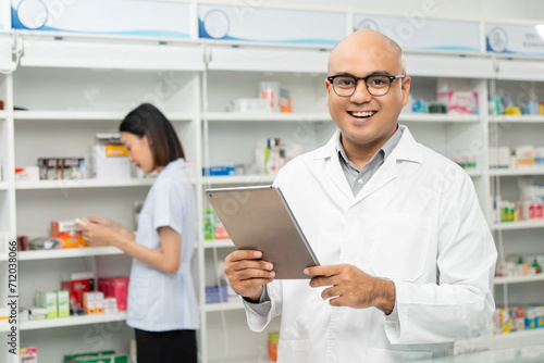 Professional asian man pharmacist checks inventory arrangement of medicine in pharmacy drugstore. Male Pharmacist wearing uniform standing near drugs shelves counter prescription to customers