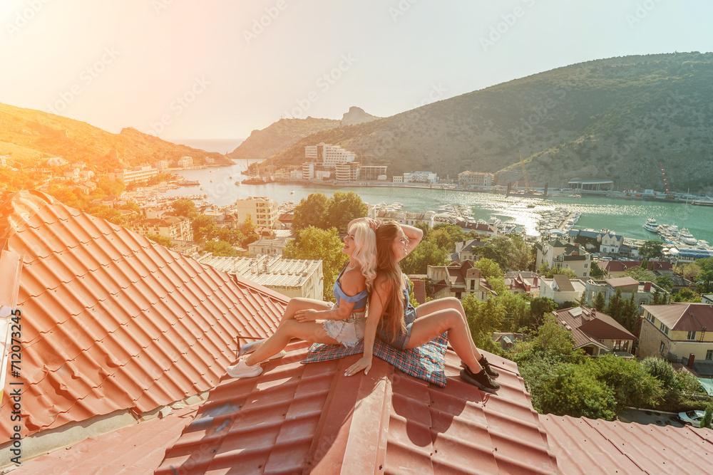 Two women sitting on a red roof, enjoying the view of the town and the sea. Rooftop vantage point. In the background, there are several boats visible on the water, adding to the picturesque scene.