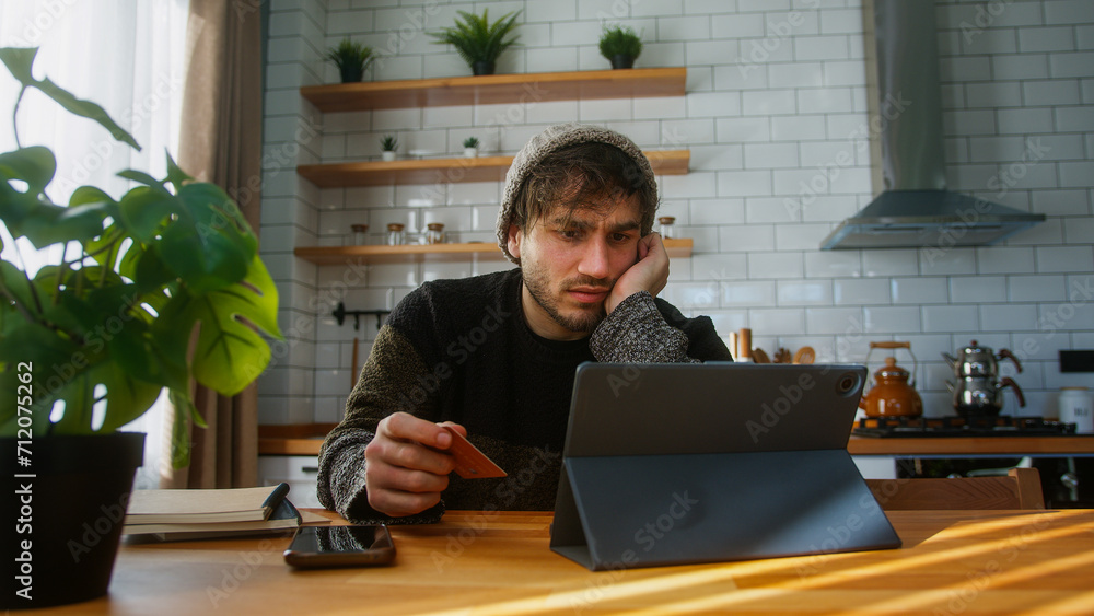 A man with beanie sitting in kitchen at home entering credit card number on tablet device for makes distant goods purchase. Credit card is declined or out of limit, feeling sad A man with beanie sitting in kitchen at home entering credit card number on tablet device for makes distant goods purchase. Credit card is declined or out of limit, feeling sad