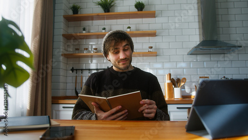 Young man student with beanie sitting in modern kitchen at home looking at the notes in notebook he is holding in his hand, reading, thinking and changing the pages	
 photo