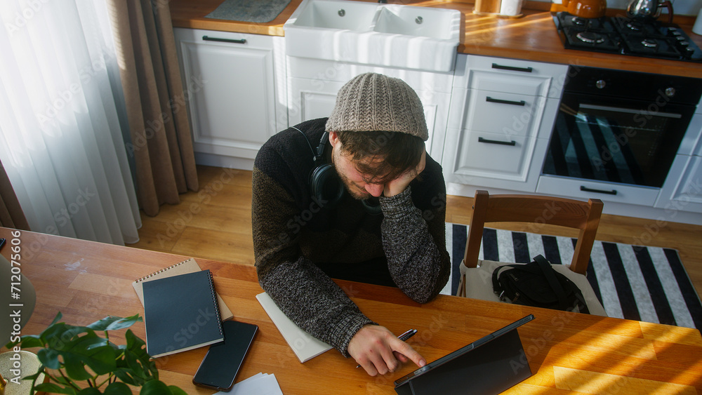 A student male with beanie sitting in modern kitchen at home works with his tablet and takes notes in notebook. High angle shot A student male with beanie sitting in modern kitchen at home works with his tablet and takes notes in notebook. High angle shot
