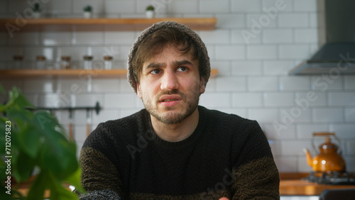 Young man with beanie sitting in modern kitchen at home looking to camera having a conference call, listening to the speaker on the video call photo