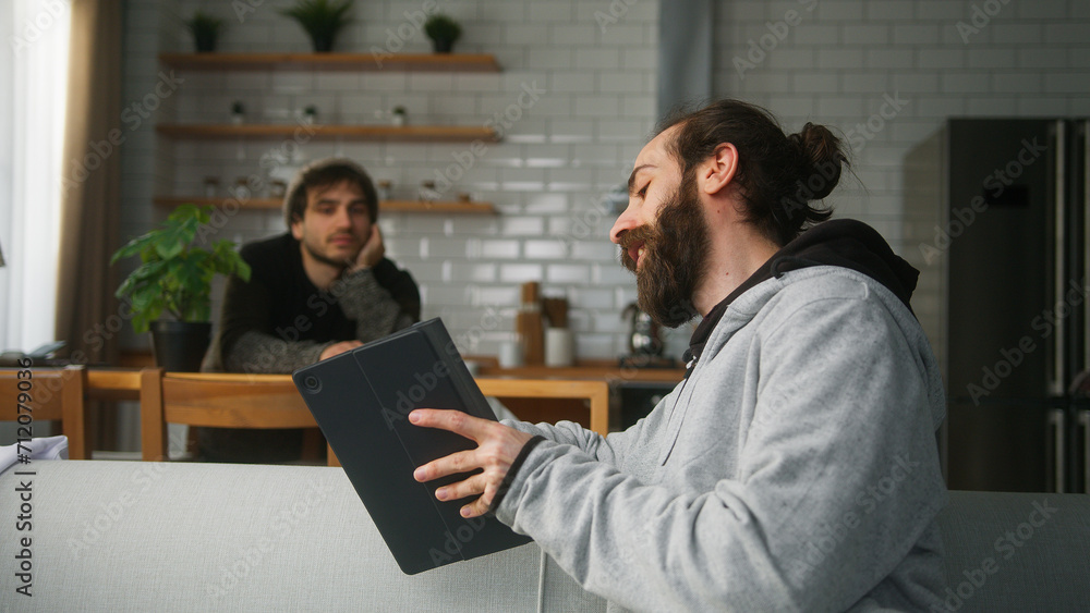 A bearded man sitting on the sofa talking to his friend sitting in the kitchen at home, showing something on his tablet computer A bearded man sitting on the sofa talking to his friend sitting in the kitchen at home, showing something on his tablet computer