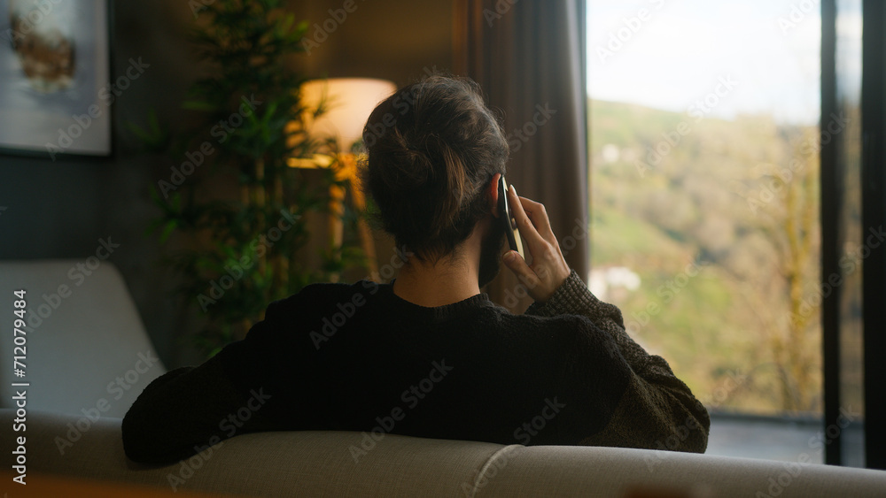 Back view of bearded man with his hair in a bun talking on his mobile phone, sitting on the sofa at home against a green landscape
Back view of bearded man with his hair in a bun talking on his mobile phone, sitting on the sofa at home against a green landscape