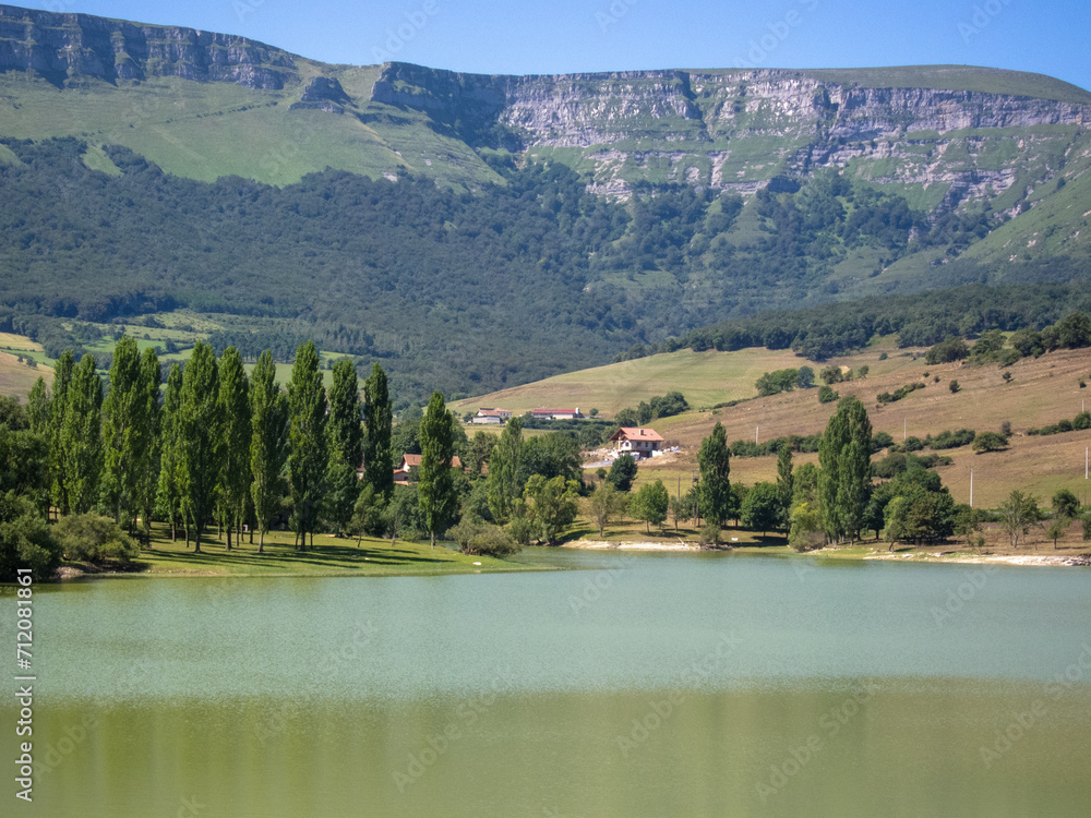 Panoramic view of the Maroño reservoir in Alava surrounded by many trees and vegetation and mountains, in the background some small rural style houses on a sunny day.
