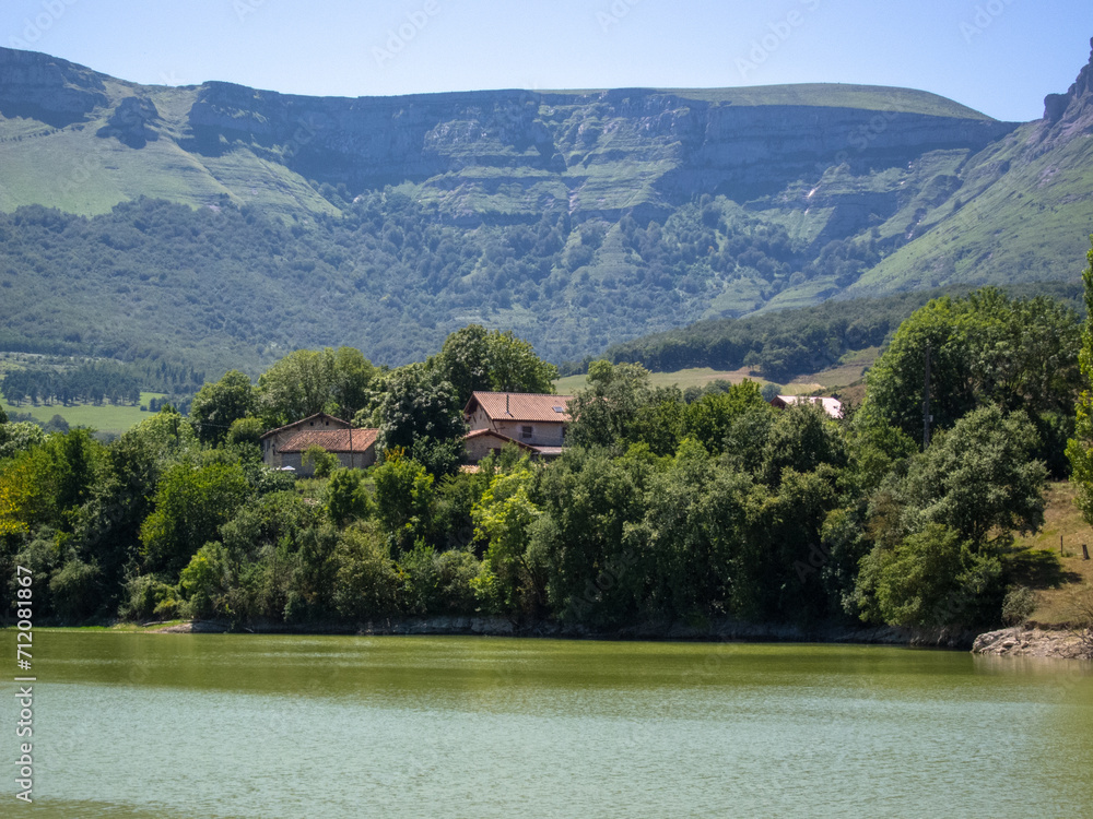 Panoramic view of the natural area of the Maroño reservoir in Alava on a sunny day with a high water level surrounded by trees, vegetation and mountains, in the background a small house.