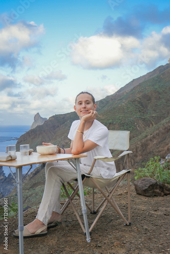 Vertical Shot: Happy Young Man in White, looking to camera and Enjoying a Cliffside Picnic in camping table and chairs, Overlooking Ocean, Late Afternoon