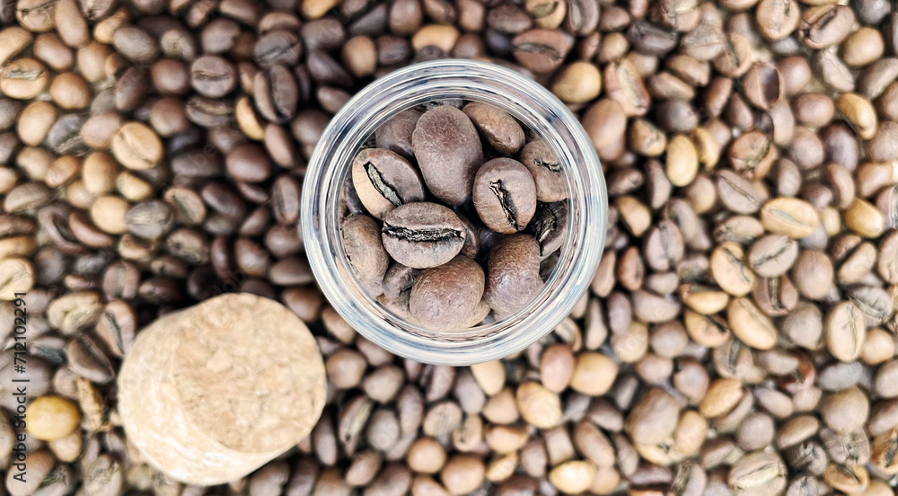 Naklejka premium Coffee beans in a small glass jar with a cork lid on the table. Coffee beans packed in a transparent, airtight storage container. Coffee seeds inside a glass jar. Flat layout.