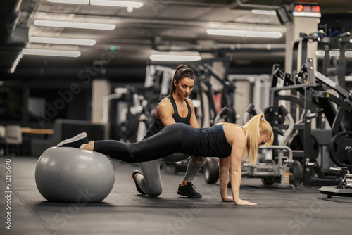 A female coach is helping a sportswoman to do planks on a fitness ball in a gym.