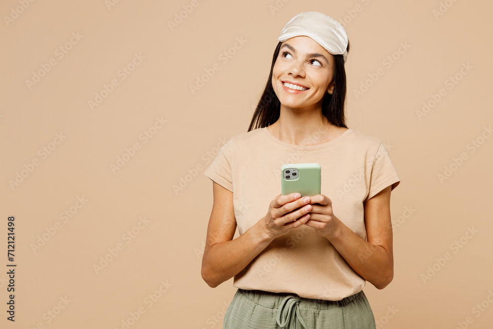 Young calm Latin woman she wear pyjamas jam sleep eye mask rest relax at home hold use mobile cell phone look aside isolated on plain pastel light beige background studio. Good mood night nap concept Young calm Latin woman she wear pyjamas jam sleep eye mask rest relax at home hold use mobile cell phone look aside isolated on plain pastel light beige background studio. Good mood night nap concept