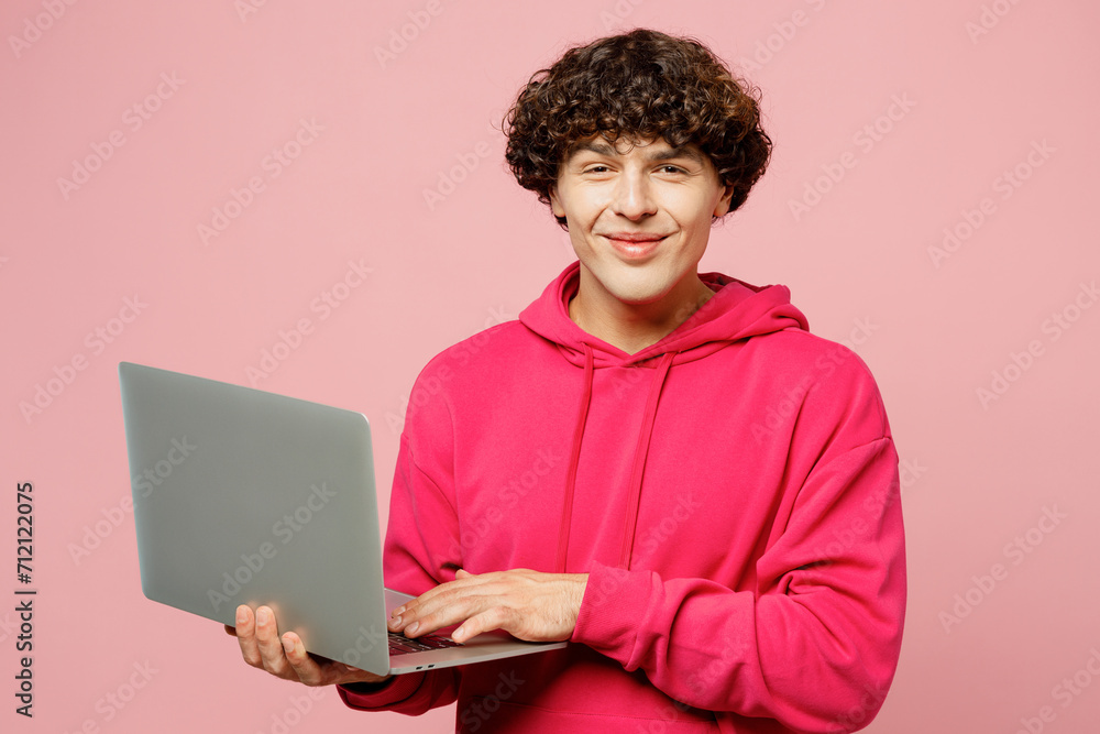 Young smiling happy Caucasian IT man he wearing hoody casual clothes hold use work on laptop pc computer look camera isolated on plain pastel light pink background studio portrait. Lifestyle concept. Young smiling happy Caucasian IT man he wearing hoody casual clothes hold use work on laptop pc computer look camera isolated on plain pastel light pink background studio portrait. Lifestyle concept.