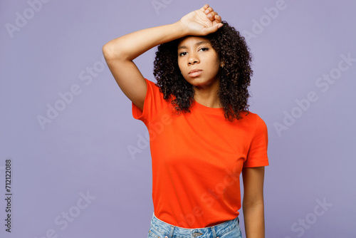 Little tired ill sick kid teen girl of African American ethnicity wear orange t-shirt put hand on forehead suffer from headache isolated on plain pastel purple background. Childhood lifestyle concept