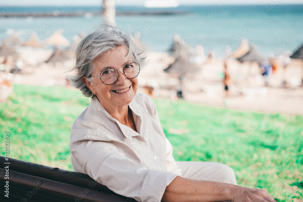 Happy relaxed senior woman with eyeglasses sitting outdoor in a bench looking at camera. Beach and sea on background
