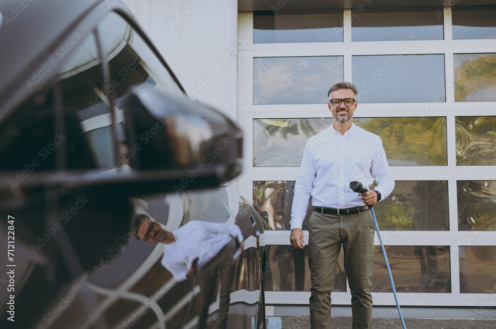 Adult cheerful businessman man wear classic shirt hold in hand electric car charger connect to electric car auto at charging station outdoor. Green energy future solution for automobile fuel concept.