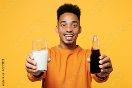 Young fun man wear orange sweatshirt casual clothes isolated on plain yellow hold give bottle of soda pop glass of milk background studio. Proper nutrition healthy fast food unhealthy choice concept. photo